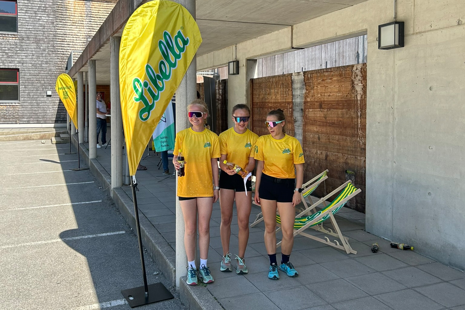 Drei Nachwuchssportlerinnen in Libella-Shirts vor Werbefahne im Stadionbereich der Chiemgau Arena.